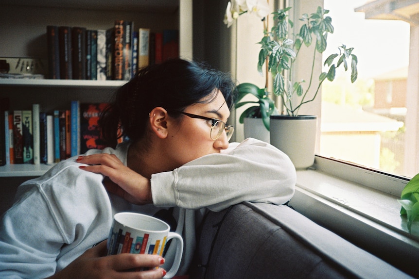 A woman with glasses and dark hair stares out the window while she drinks a coffee.