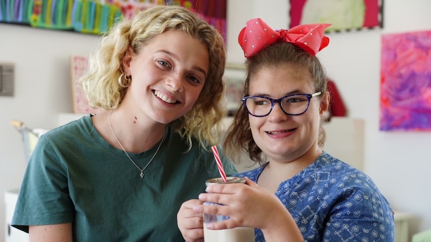 Two young women smile and pose for a photo while one sips a milky drink