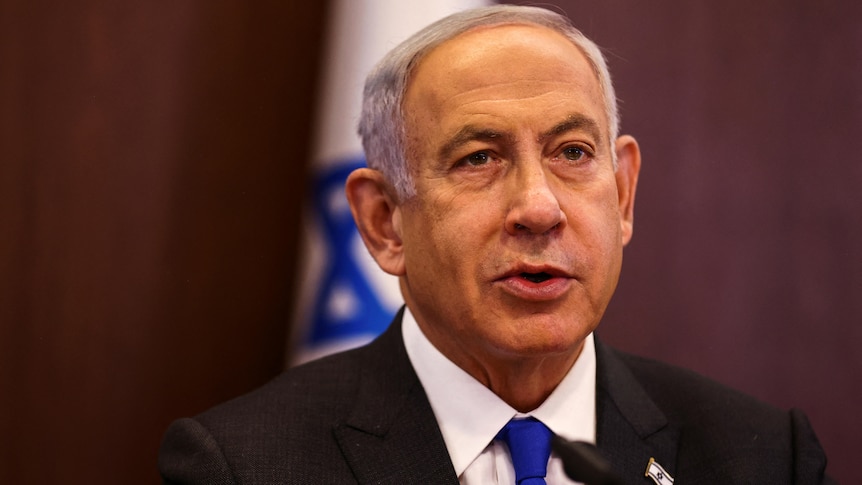 A man wearing a suit sits in front of a blue and white flag. 