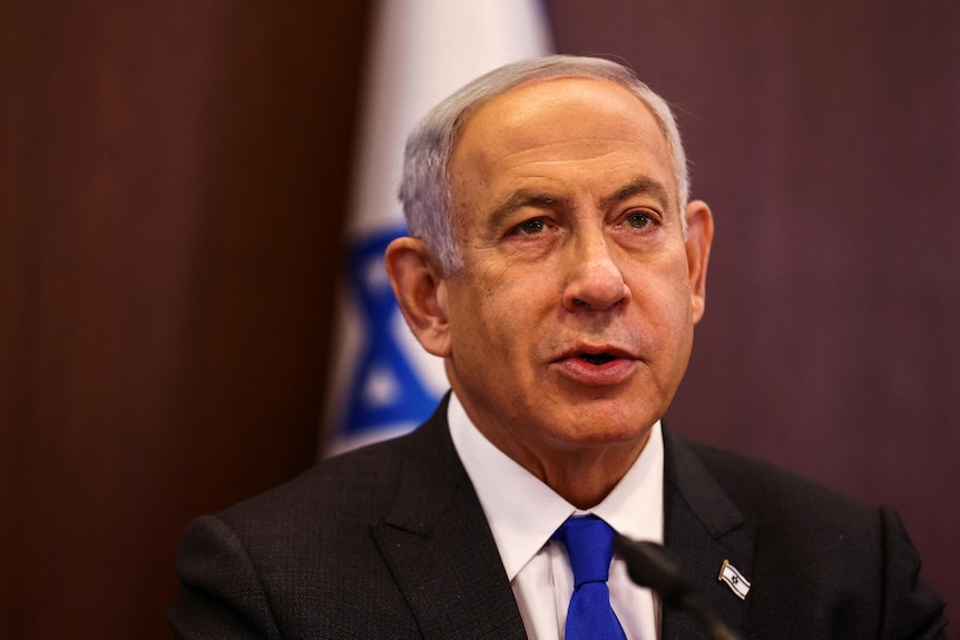 A man wearing a suit sits in front of a blue and white flag. 