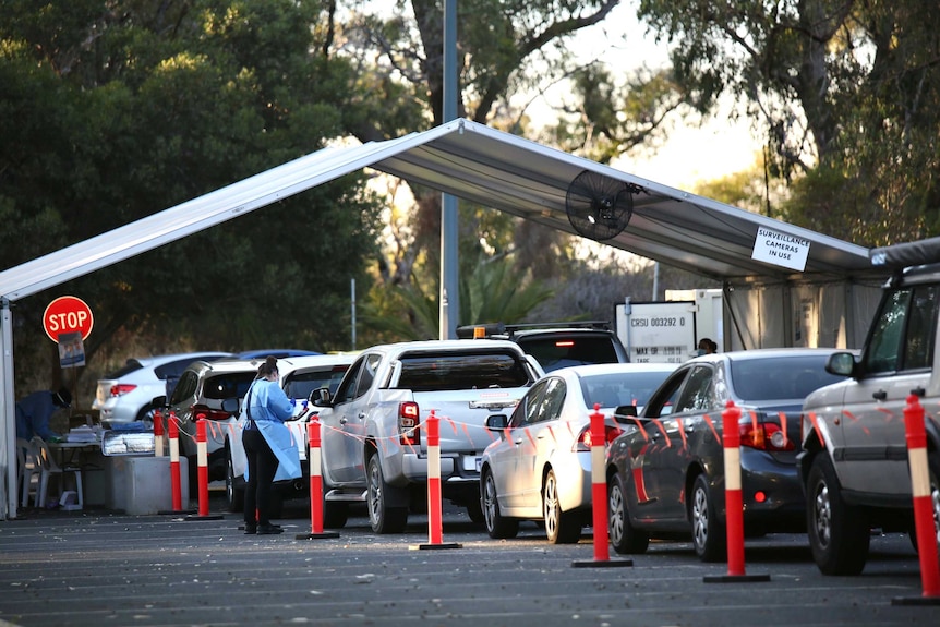 A line of cars stretches to a temporary shelter, where a woman in PPE holds a clipboard.