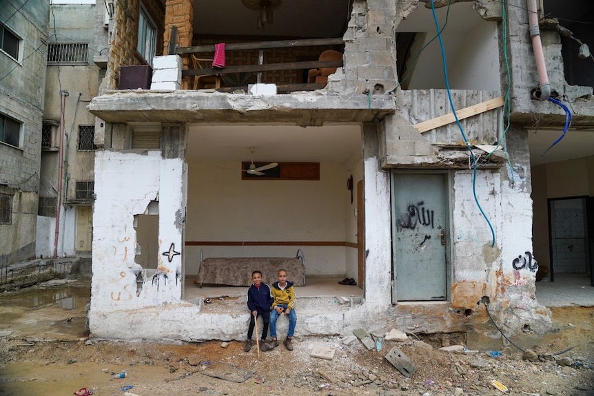 Two young boys sitting in a large hole in an exterior wall of a building. A bed can be seen in the room behind them