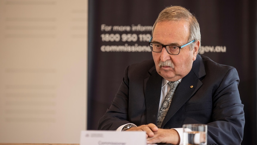 Older man with glasses sits at table, addressing the media 