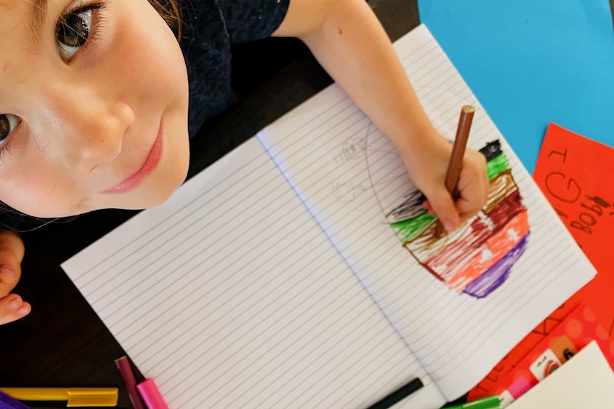 A child looks up at the camera while drawing a picture in a book.