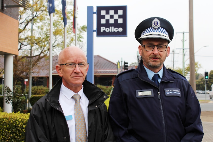 Two middle-aged policemen stand outside a police station on an overcast day. One is plain clothes, the other full uniform.