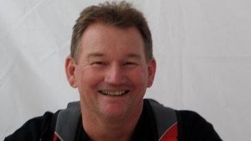 A man in a dark coloured polo shirt with red sections smiling at the camera.