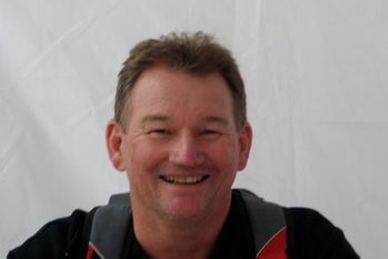 A man in a dark coloured polo shirt with red sections smiling at the camera.
