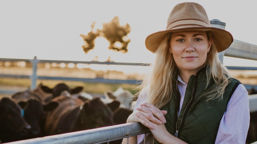 A woman in a brimmed hat, long-sleeve shirt and vest stands leaning on a cattle fence, cattle behind her