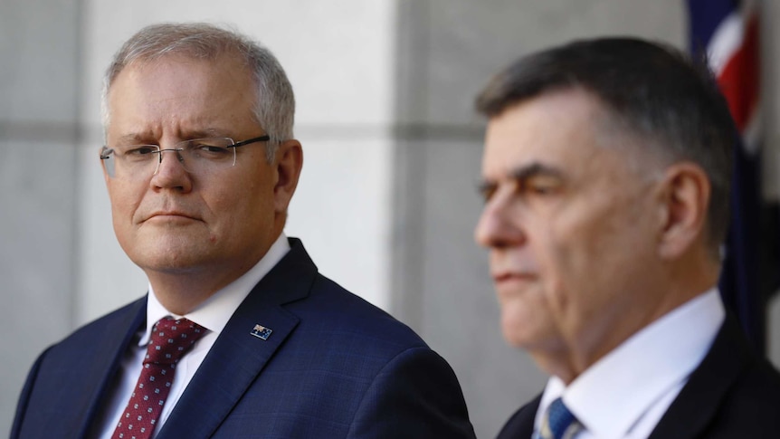 Scott Morrison looks at Brendan Murphy as he speaks at a podium.