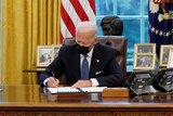 Joe Biden in the Oval Office signs a document while sitting at a desk while a man in a suit stands to his left.