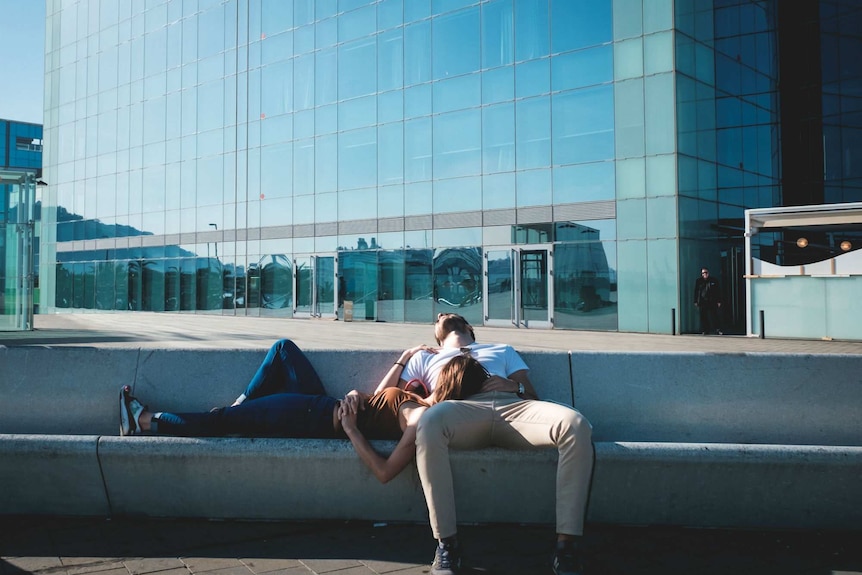 A man and a woman nap on a concrete bench in front of an office building.