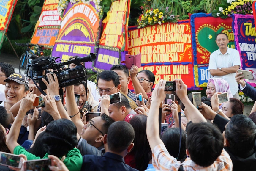 Ahok is guided through a crowd by his handlers near the wall of floral tributes