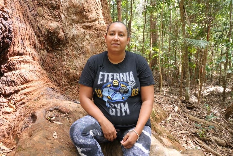 Aboriginal woman sits on large root at the base of a giant Blackbutt tree.