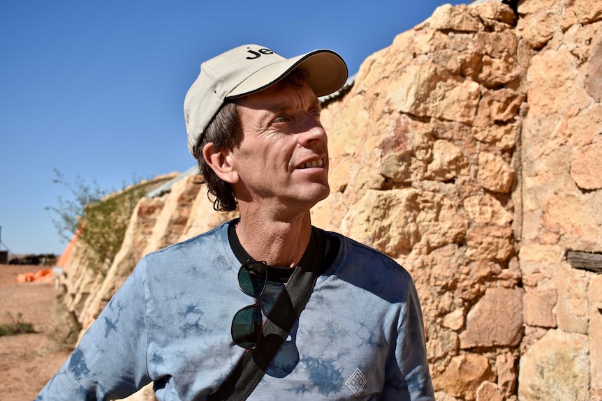 A man in a tan coloured cap looks at a stone shed on a bright sunny day.