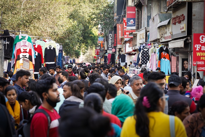 Crowds of people gather on a street with shop fronts on one side and open market stalls on the other