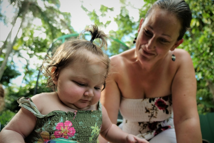 An infant plays on a mat outside as a mum watches.