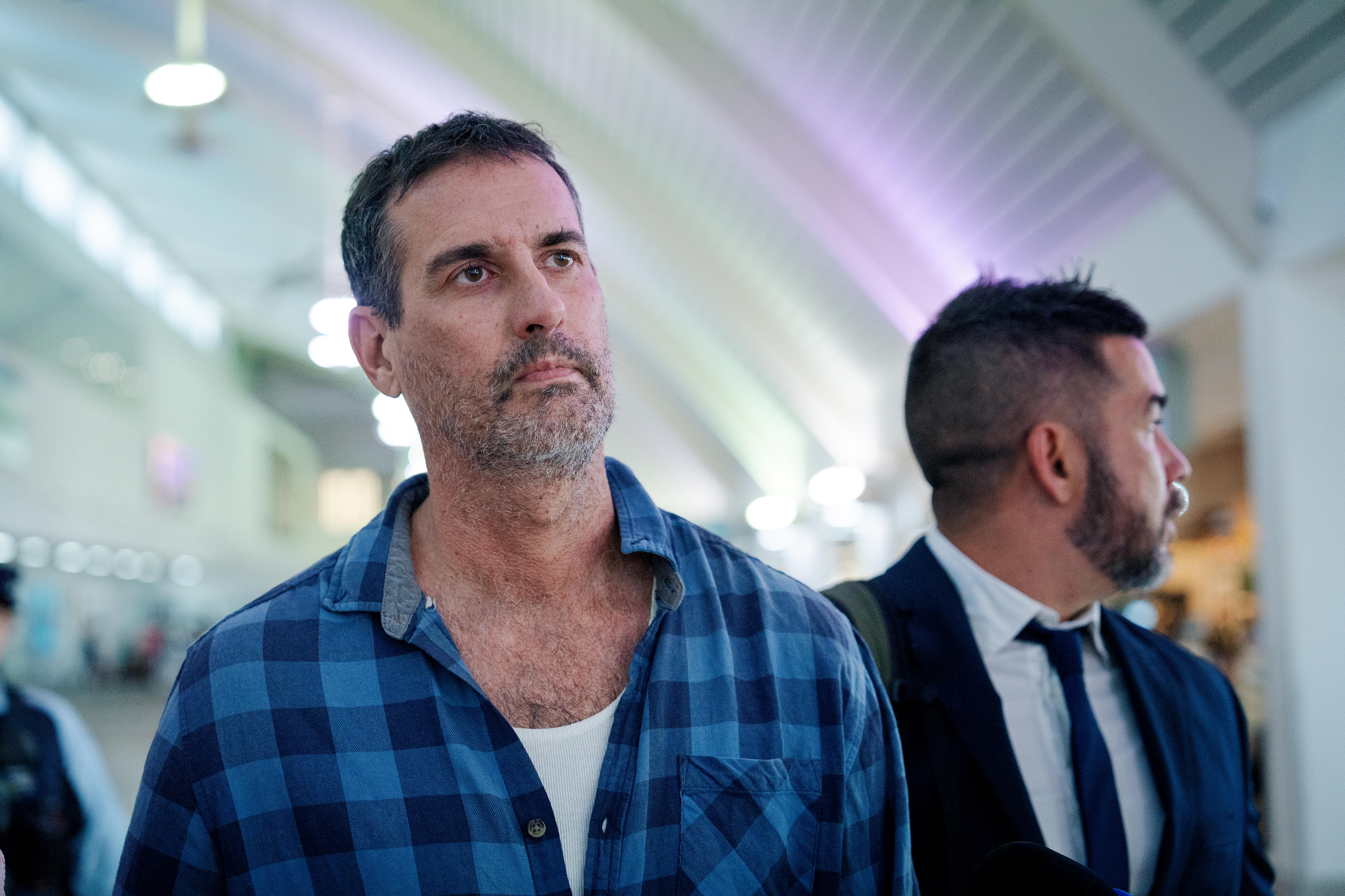 Un hombre mira a lo lejos mientras camina por un aeropuerto, flanqueado por un policía a un lado.