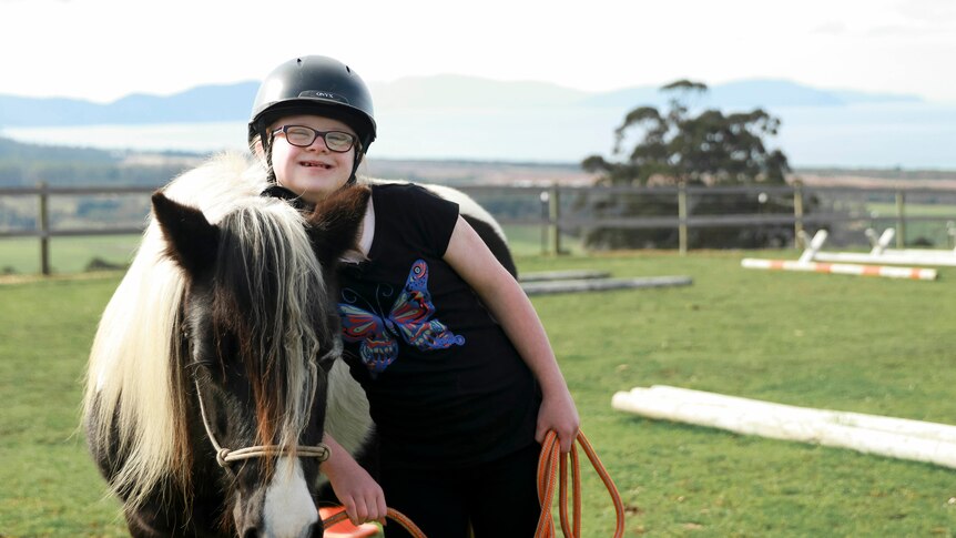 Young girl leans on pony at riding arena surrounded by rolling green hills.