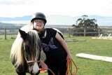 Young girl leans on pony at riding arena surrounded by rolling green hills.