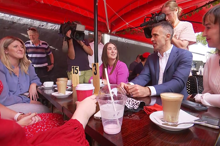 A man has coffee with three women with two TV cameras looking on