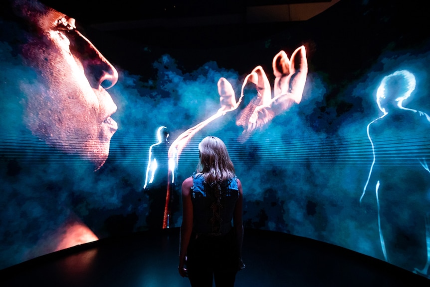 A person stands in front of a large screen with side on view of person with hand raised and surrounded by smoke.