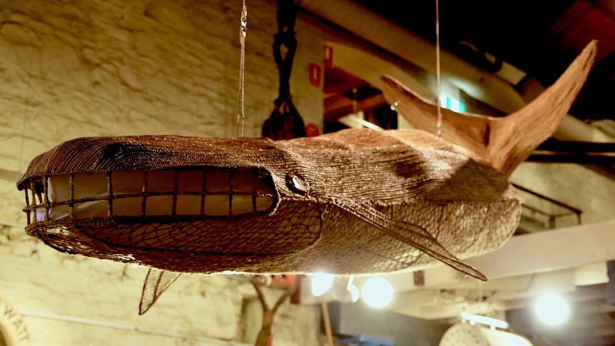A woven sculpture of a whale suspended near a boat in a museum.