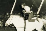 ABC cameraman Warwick Curtis hangs from a suspension cord as he rides a horse at the circus.