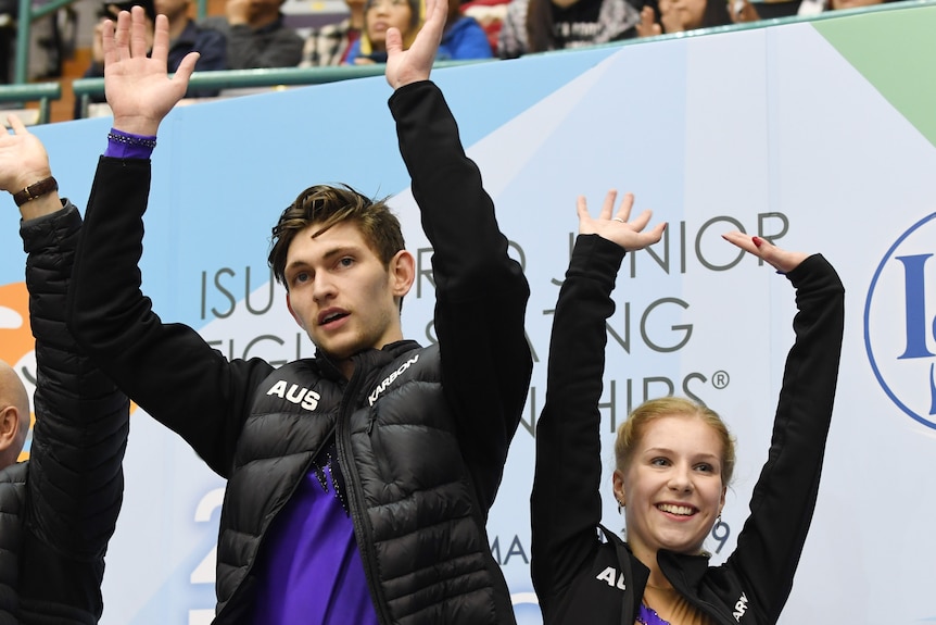 An Australian figure skating pair stand with their coach smiling with arms raised acknowleding the crowd after a win.
