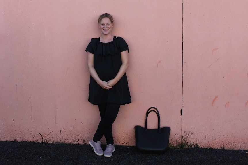 A young woman stands leaning against a pink wall