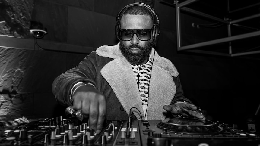 Madlib wearing sunglasses and headphone twiddling a knowb as he DJs a live set.