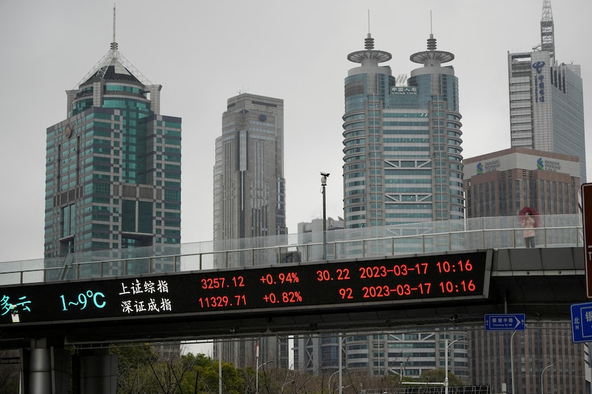 An electronic board on the side of a pedestrian bridge with red stock market figures. High rise buildings are in the background.