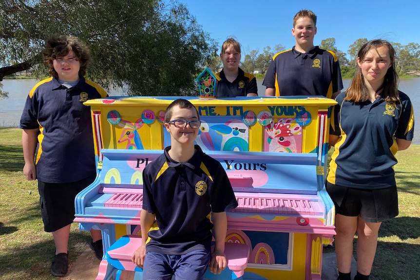 A group of children stand around a brightly-coloured piano which is placed alongside a river. 