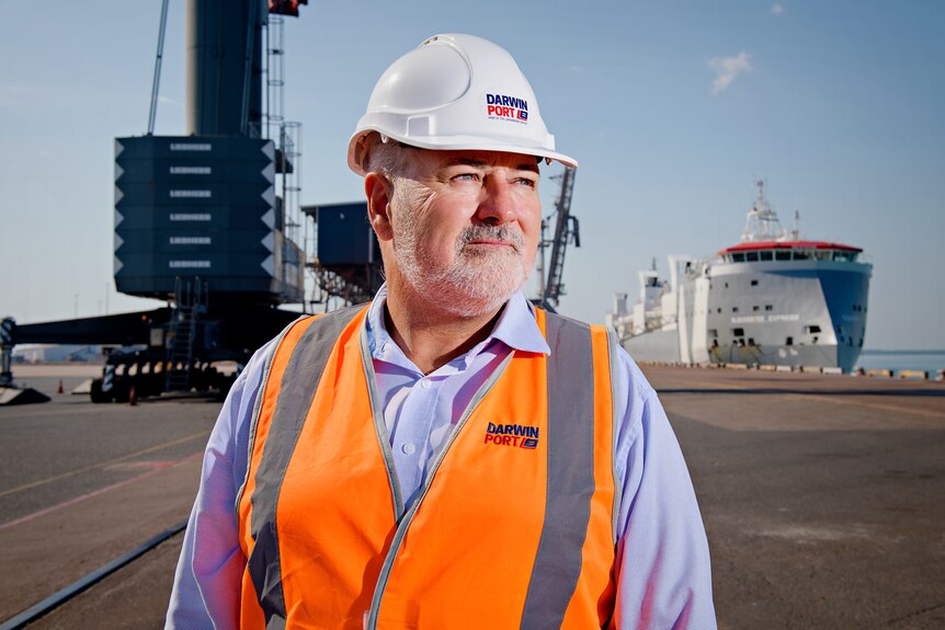 A serious-looking man in a hard hat and high vis vest standing with his hands on his hips, with port infrastructure behind him.
