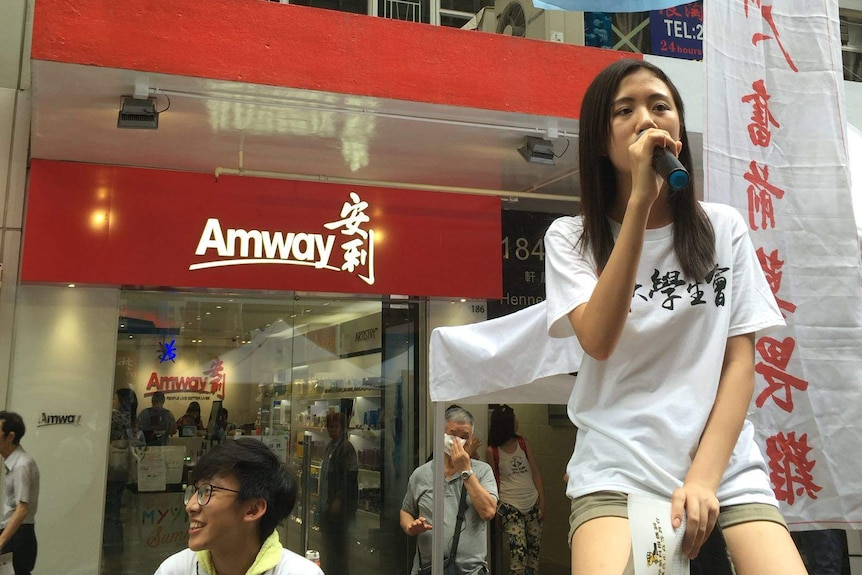 Ms Cheung as a protest. She speaks through a microphone in front of blue and white flags.