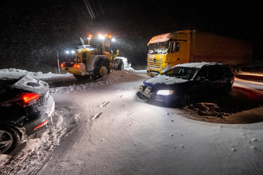 A snow plow clears snow from a road as cars and trucks are stuck on a road at night