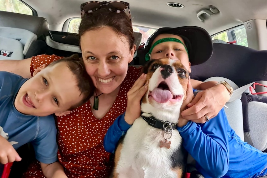 a woman with her arms and around her two young sons in the back of a car, with a dog on their laps