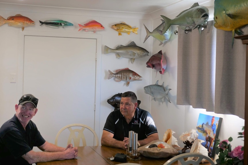 Two blokes sitting at a table in front of a wall of fish. 
