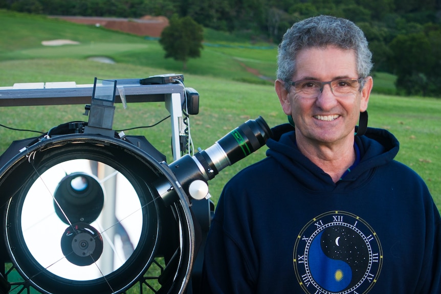A grey haired man with glasses stands next to a big telescope