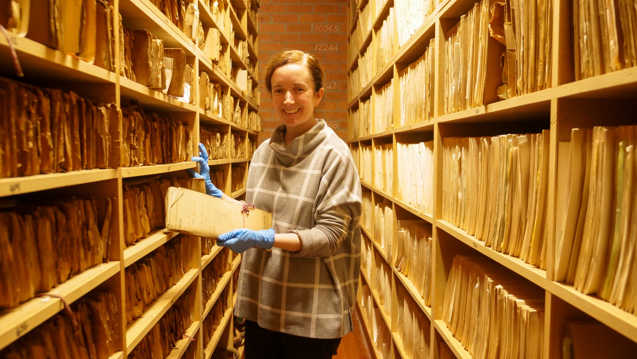 A woman stands in a tight corridor surrounded by paper files. 