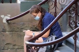 A woman sitting on steps and wearing a face mask looks at her mobile phone.