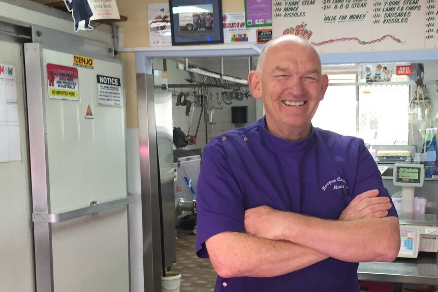 Glen Innes butcher Terry Hooper standing behind his shop's counter, smiling and with arms folded.