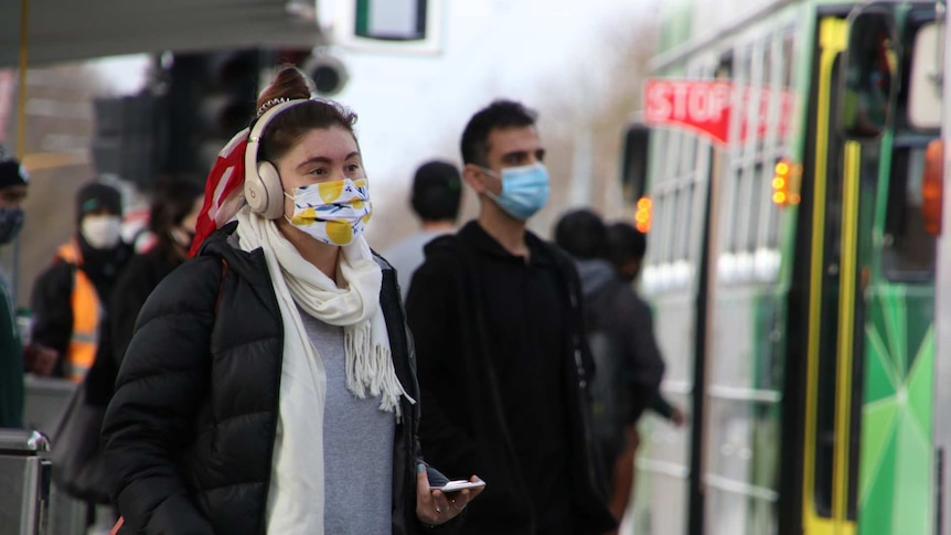 A person in a lemon-patterned face mask walks towards a tram.