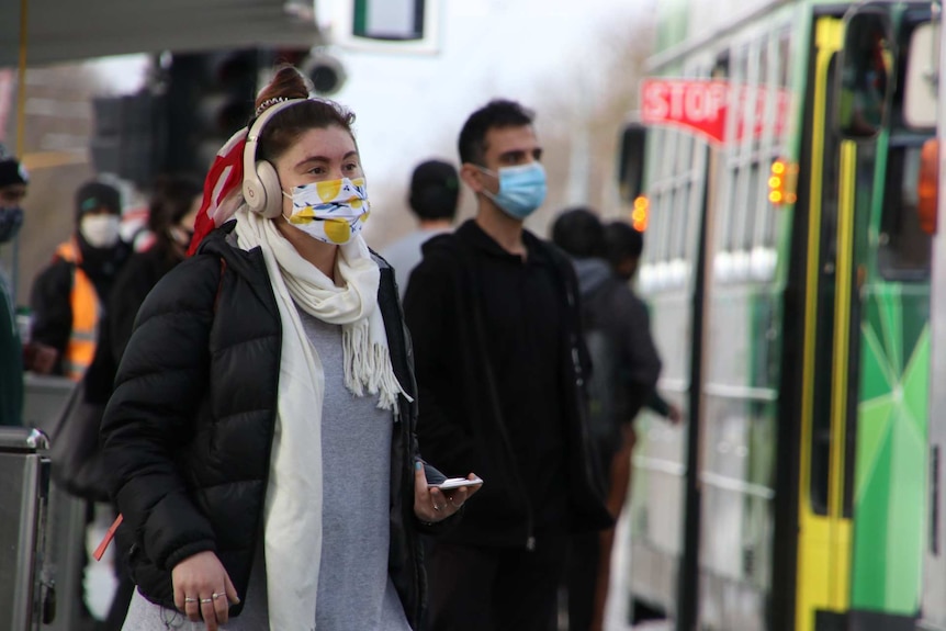 A person in a lemon-patterned face mask walks towards a tram.