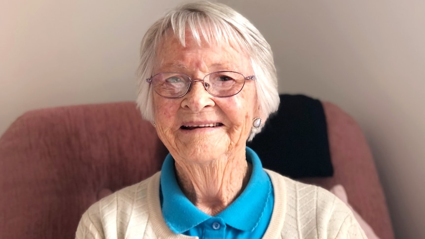 An elderly woman with glasses smiles at the camera