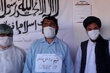 Men in surgical gowns and masks stand in front of a wall with information posters about coronavirus written in Afghan script.