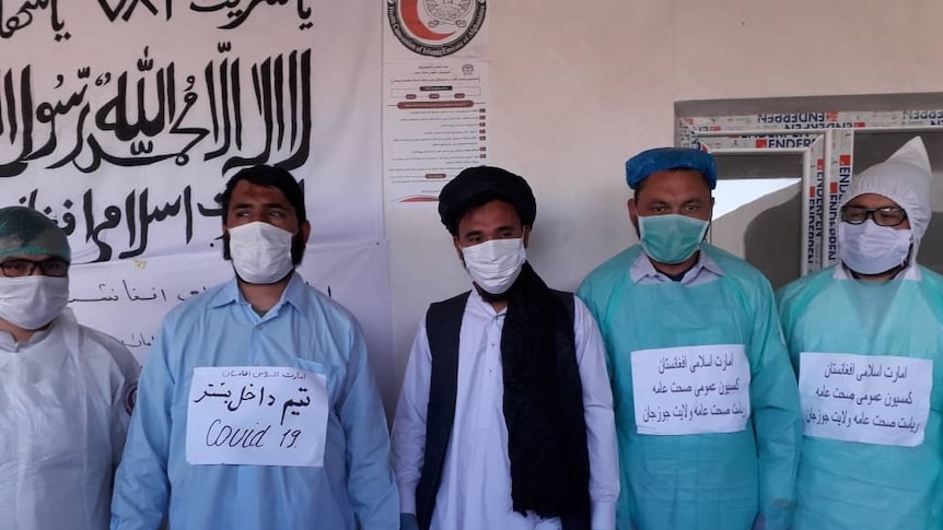 Men in surgical gowns and masks stand in front of a wall with information posters about coronavirus written in Afghan script.
