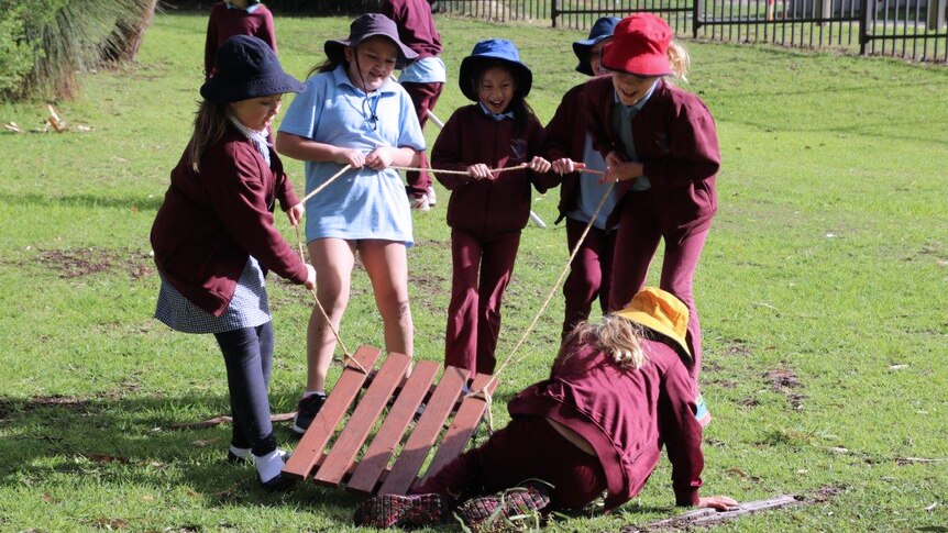 A group of five young female students stand pulling a wooden sled by a rope on grass with another girl on the ground.