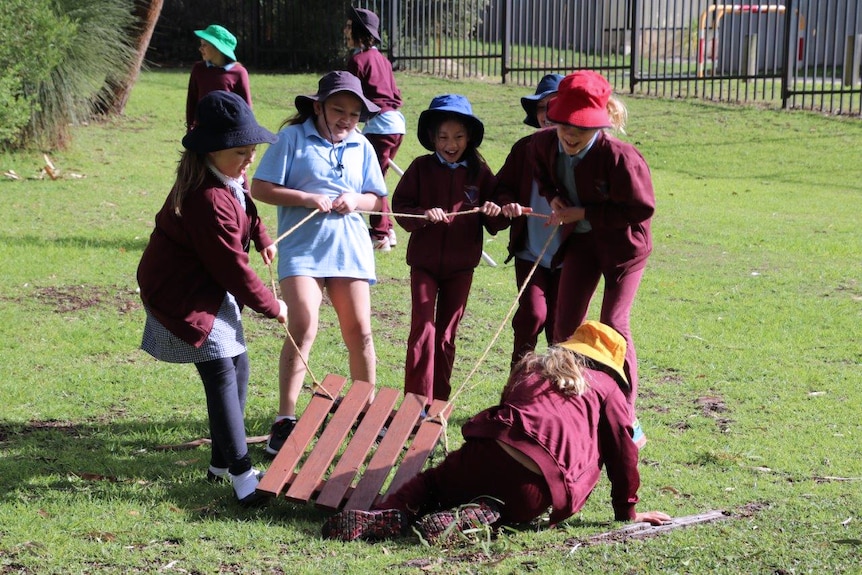 A group of five young female students stand pulling a wooden sled by a rope on grass with another girl on the ground.