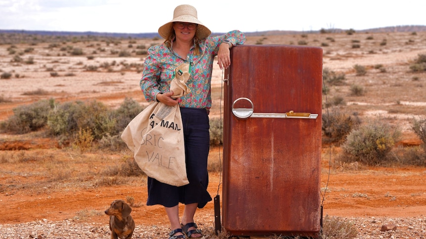 A woman wearing a hat rests her arm on top of a rusted refrigerator while a small dog waits next to her.