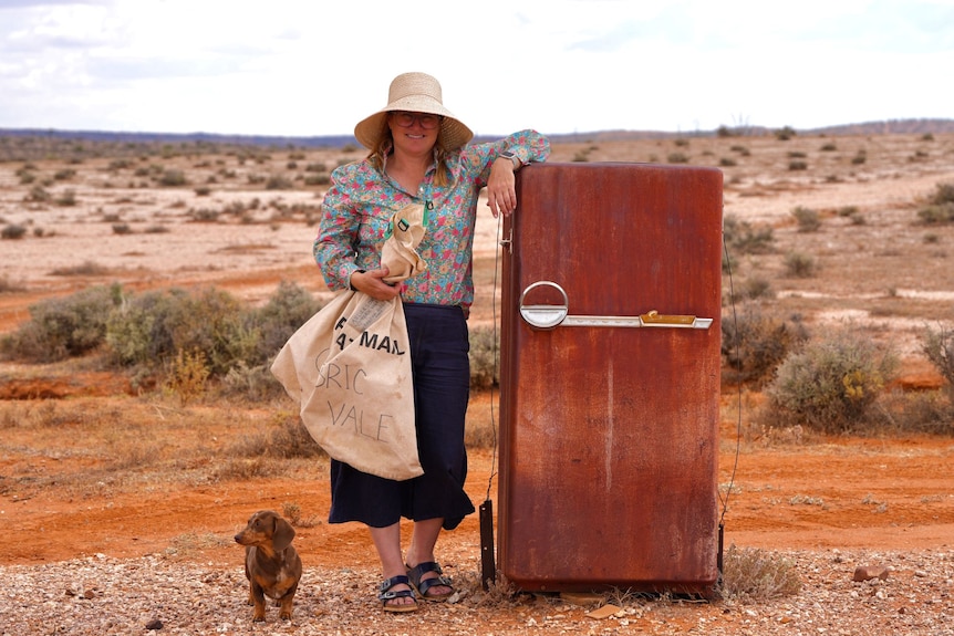 A woman wearing a hat rests her arm on top of a rusted refrigerator while a small dog waits next to her.